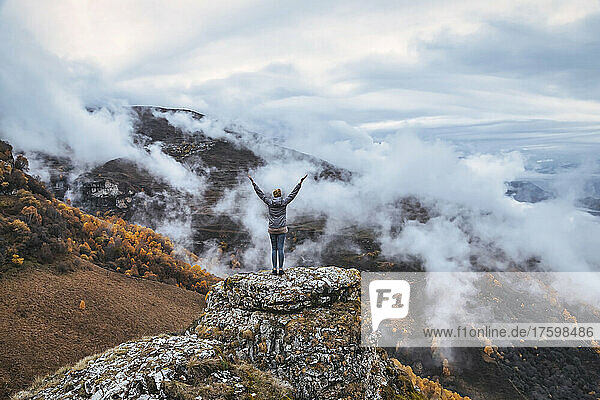 Female hiker standing with raised arms on top of outcrop in autumn mountains of North Caucasus
