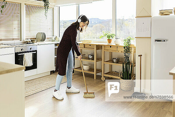 Young woman cleaning kitchen floor with broom at home