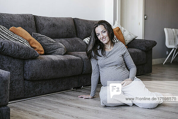 Lächelnde schwangere Frau sitzt zu Hause am Sofa