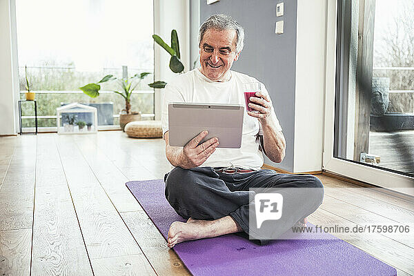 Smiling senior man sitting cross-legged on exercise mat watching video through tablet PC at home