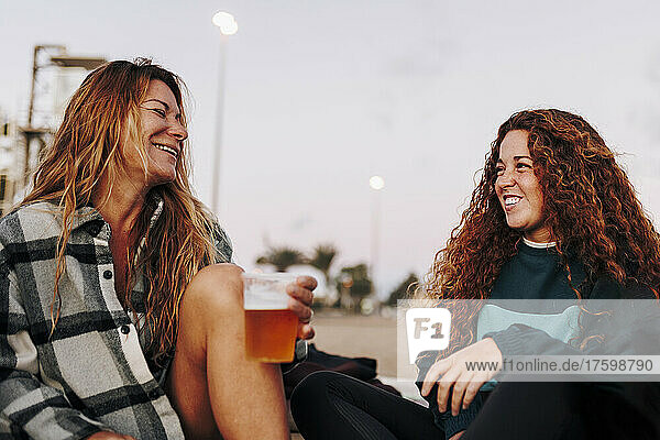 Glückliche Frau hält einen Bierbecher in der Hand und unterhält sich mit einer Freundin am Strand
