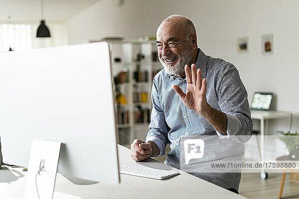 Cheerful senior businessman waving hand on video call through desktop computer in home office