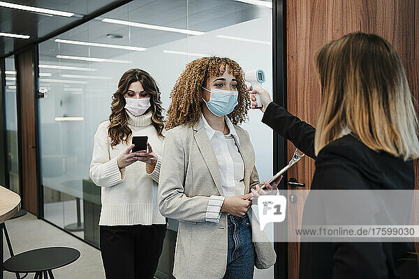 Businesswoman checking temperature of colleague wearing protective face mask in office