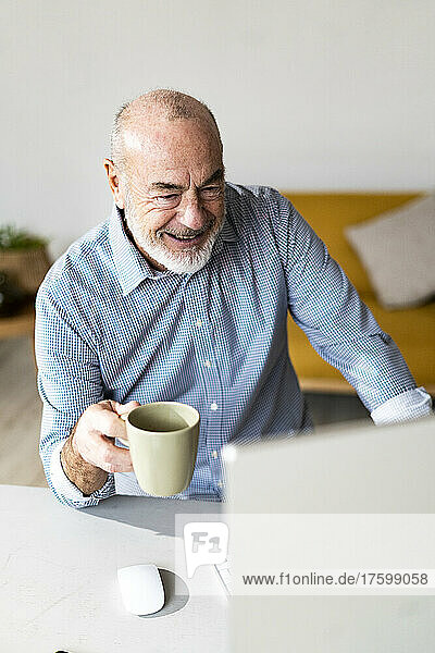 Happy senior freelancer holding coffee mug working in home office