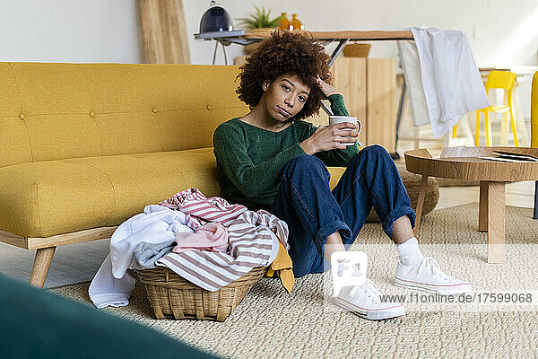 Tired Afro woman with hand in hair holding coffee mug sitting by basket at home