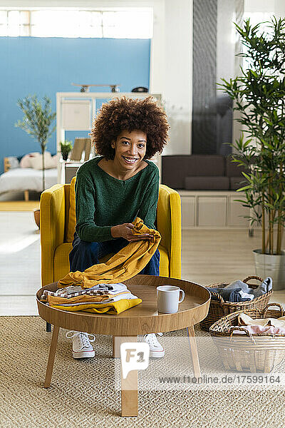 Smiling young woman with curly hair folding clothes sitting on chair in living room at home
