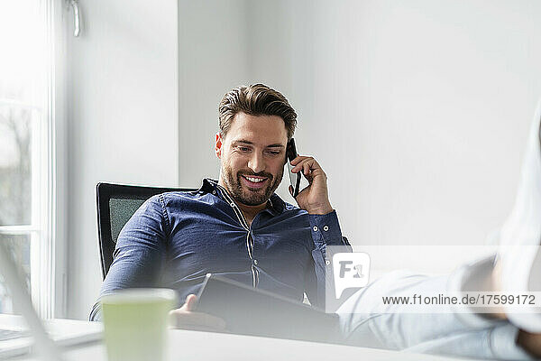 Smiling businessman with tablet PC talking on smart phone in office