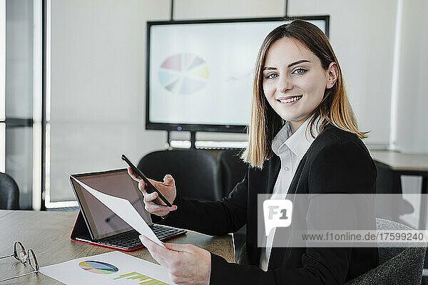 Smiling working woman holding smart phone and document at desk in office