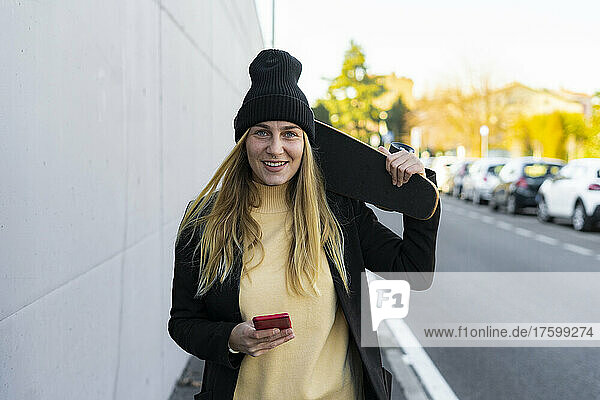 Smiling woman with smart phone and skateboard on street