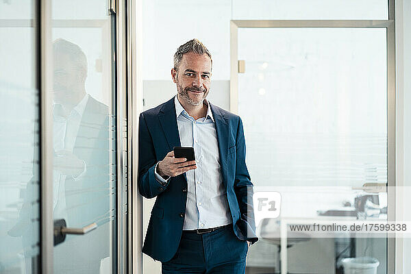 Smiling businessman leaning with hand in pocket holding smart phone at office