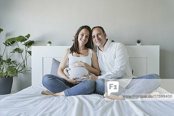 Glücklicher Mann sitzt mit schwangerer Frau zu Hause im Bett