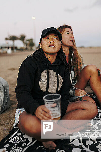 Thoughtful woman with beer cup sitting by friend at beach