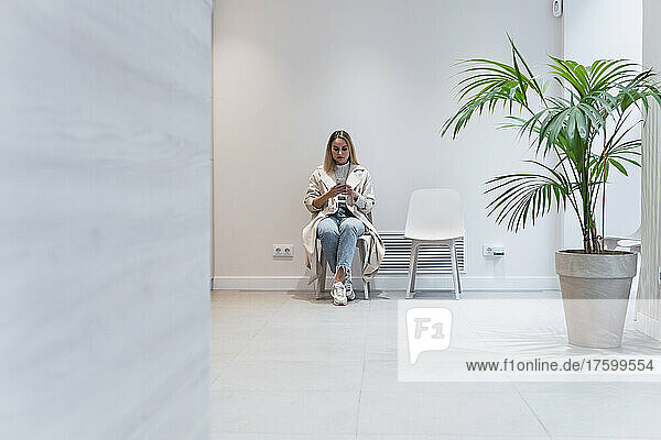 Young woman using mobile phone sitting in waiting room at clinic