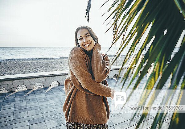 Smiling young woman wearing sweater by palm leaf at promenade