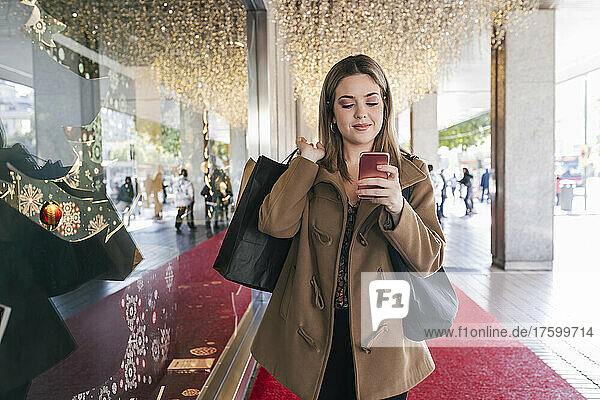 Smiling with shopping bags using mobile phone by store at Christmas