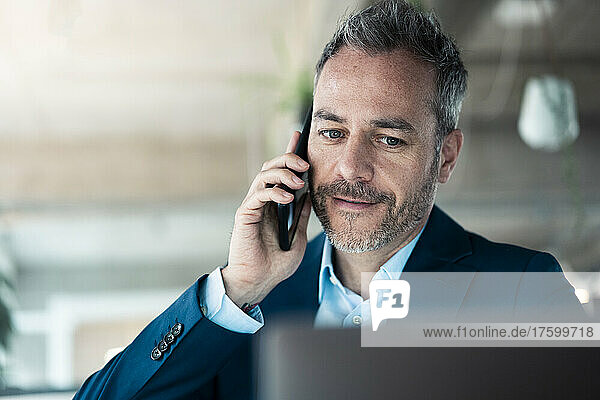 Businessman talking on mobile phone looking at laptop in workplace
