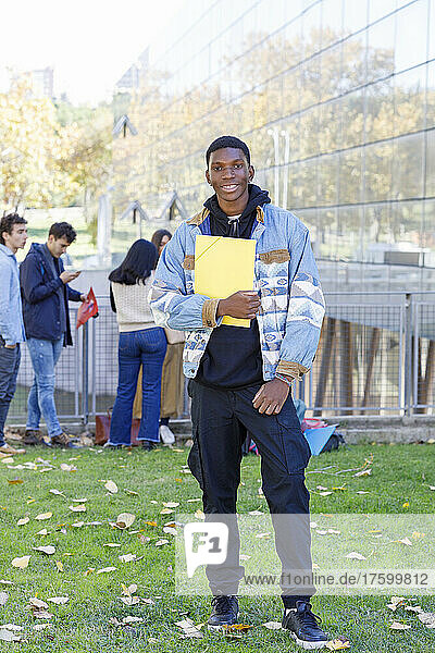 Smiling teenager holding file standing with friends in background at campus