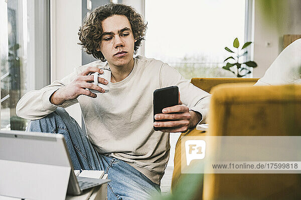 Young man holding coffee cup using smart phone in living room