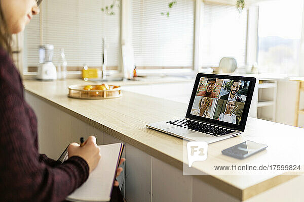 Young working woman with notebook doing video call with colleagues on laptop at home