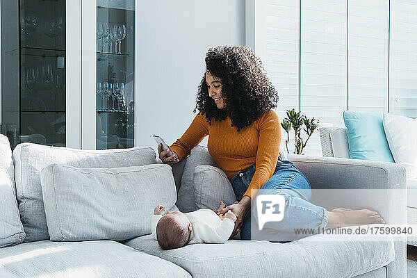 Mother using smart phone sitting by baby boy on sofa at home