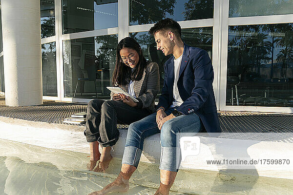 Gemischtrassige Geschäftskollegen teilen sich einen Tablet-PC und sitzen mit Beinen im Pool