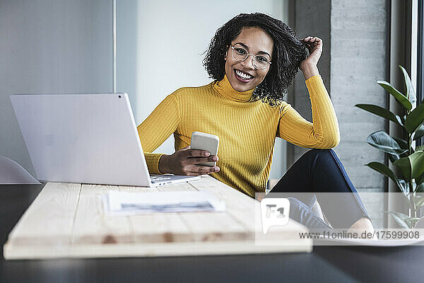 Happy businesswoman with mobile phone by laptop at work place