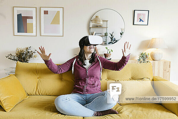 Happy woman wearing virtual reality headset gesturing on sofa at home