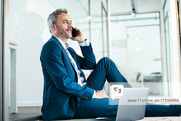 Smiling businessman sitting with laptop talking on mobile phone in office corridor
