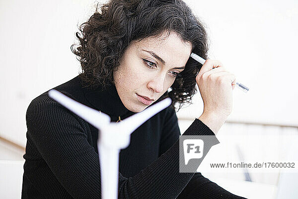 Nachdenkliche junge Geschäftsfrau hält Stift neben Windturbinenmodell im Heimbüro