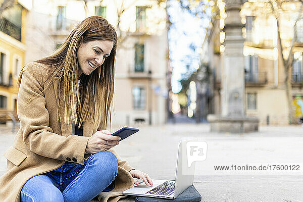 Lächelnde Frau mit Laptop  die über ihr Mobiltelefon im Internet surft  sitzt auf der Stadtstraße