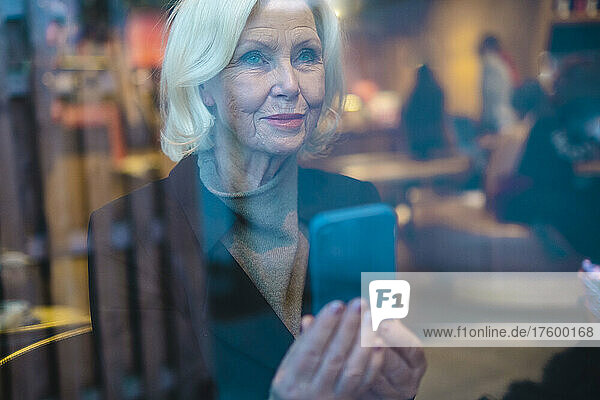 Senior woman with smart phone seen through window