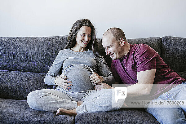Pregnant woman holding headphones over belly looking at man sitting on sofa