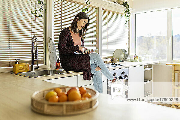 Frau sitzt zu Hause auf der Küchentheke und benutzt einen Tablet-PC