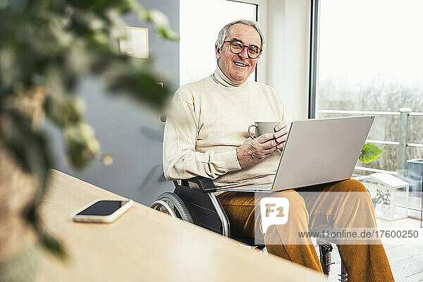 Smiling disabled senior man holding coffee cup sitting with laptop on wheelchair at home