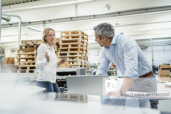 Smiling blond businesswoman sharing ideas with colleague leaning on desk in factory