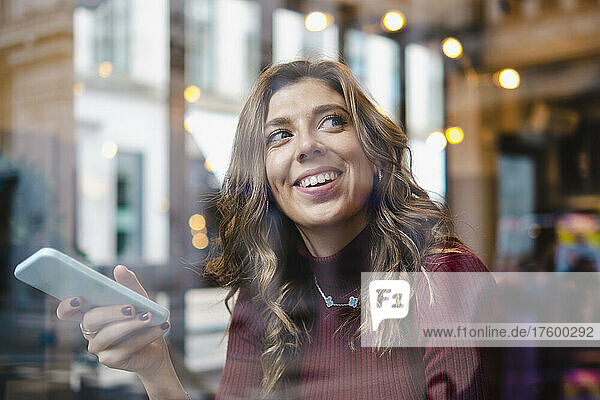 Happy woman with smart phone seen through window of cafe