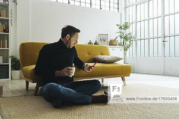 Mature man with coffee mug using smart phone in living room at home