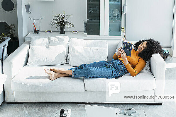 Woman reading book lying on sofa in living room at home