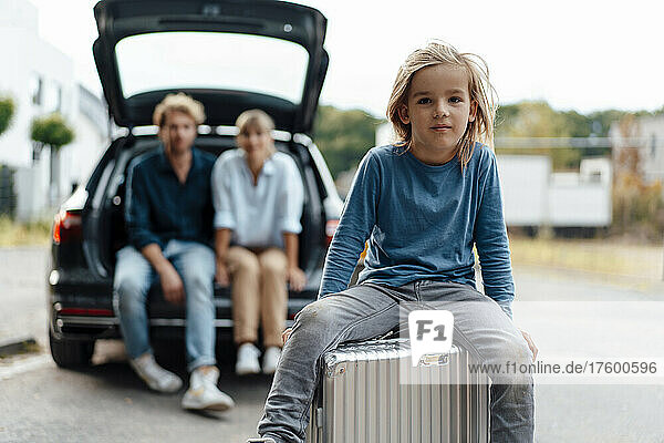 Junge sitzt auf Koffer mit Eltern im Hintergrund im Urlaub