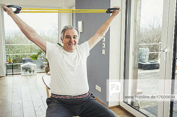 Smiling senior man with arms raised practicing with resistance band at home
