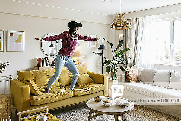 Young woman wearing virtual reality headset standing with arms outstretched on sofa in living room