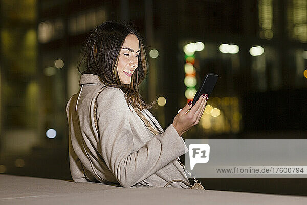 Young woman using smart phone leaning on wall at night