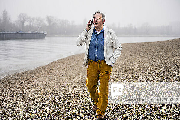 Glücklicher älterer Mann  der mit dem Smartphone spricht und mit der Hand in der Tasche am Strand läuft