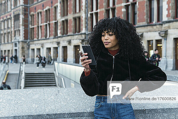 Fashionable young curly haired woman using smart phone in city