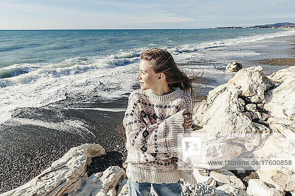 Smiling teenage girl in sweater looking at sea at beach on sunny day