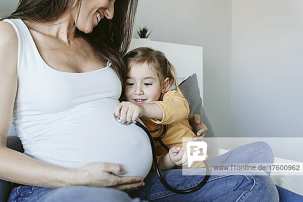 Smiling pregnant woman looking at daughter listening through stethoscope