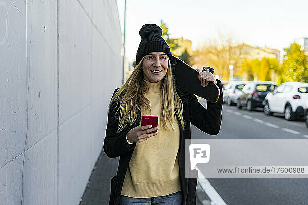 Blond woman with smart phone and skateboard by wall on street