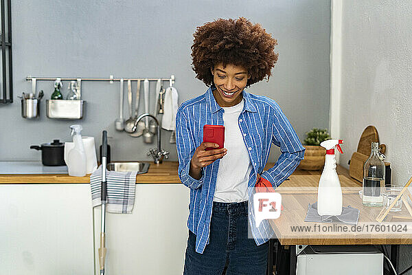 Smiling young Afro woman text messaging on smart phone by spray bottle at kitchen counter