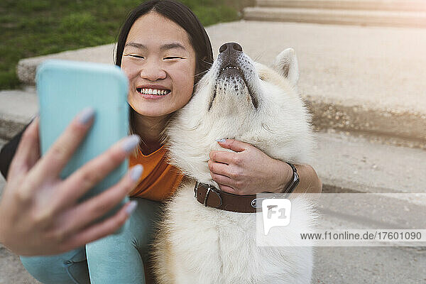 Smiling woman taking selfie with Akita dog on smart phone in park