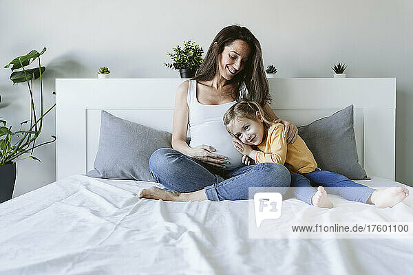 Happy pregnant woman sitting with daughter on bed at home